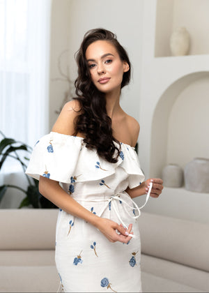 Woman wearing a white linen off-shoulder dress with floral patterns in a modern indoor setting.