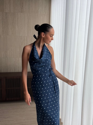 A woman wearing a navy blue halter-neck midi dress with a white polka dot pattern, standing in a modern room next to white window curtains.
