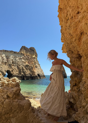 Woman in a Bohomey striped linen dress standing between two rocky cliffs with clear blue water and sky.