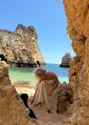 Woman in a Bohomey striped linen maxi dress sitting on a sandy beach with rocky cliffs and clear blue water.