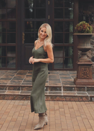 Woman in a Bohomey moss green linen slip dress standing in front of a large glass door with brick walls.