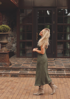 Woman in a Bohomey moss green linen slip dress standing in front of a large glass door with brick walls.