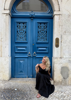 Woman in a Bohomey black linen dress sitting in front of a blue door on a stone pavement.