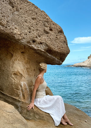 Woman in Bohomey linen dress sitting under a large rock by the ocean