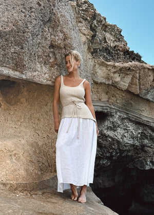 Woman in a Bohomey beige and white linen dress standing on a rocky outcrop with a cave in the background.
