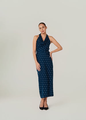 A woman modeling a sleeveless halter-neck maxi dress with a small white polka dot linen pattern on a dark navy blue base, standing against a solid light gray studio background.