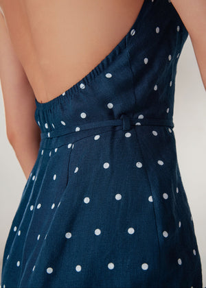 Close-up of a navy blue sleeveless dress with a white polka dot linen pattern, featuring a low open back and a tie belt at the waist, worn by a model.