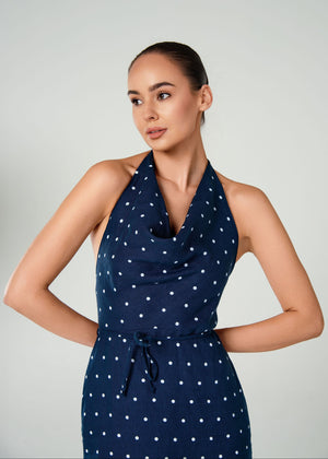 A woman modeling a navy blue halter-neck dress with a white polka dot linen pattern against a light gray studio background.