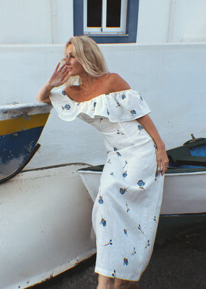 Woman in a white linen floral dress leaning against a boat near a white building with blue accents.