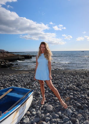 Woman in a BOHOMEY linen light blue dress standing on a rocky beach with a boat and ocean in the background.