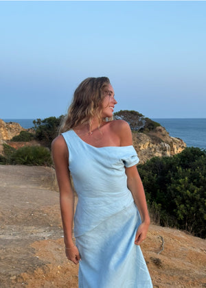 Woman in a BOHOMEY light blue linen dress standing on a coastal path with ocean and cliffs in the background