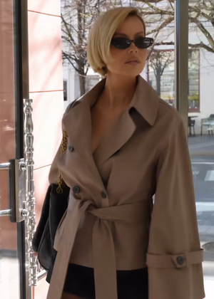 Woman in a beige trench coat and sunglasses standing in front of a glass door.