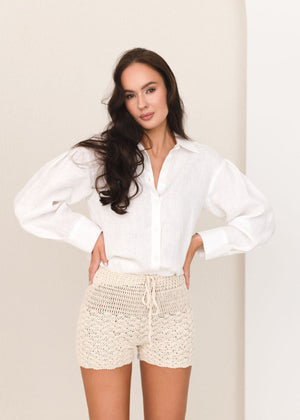 Woman wearing a white linen blouse and beige crochet shorts against a plain background