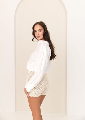 Woman wearing a white linen blouse and lace crochet shorts standing against a plain background