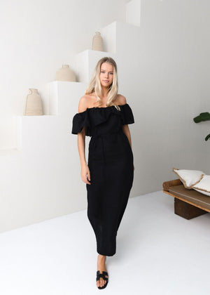 Woman in a black linen off-shoulder dress standing in a minimalistic room.
