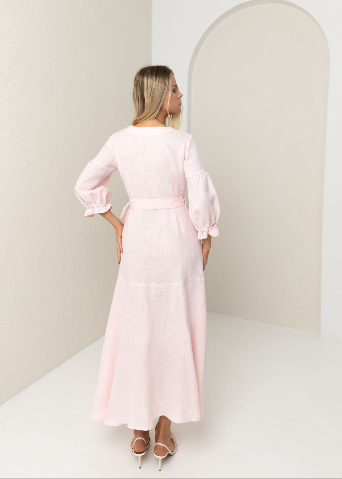 Woman wearing a light pink linen dress standing in a minimalistic room with a white wall and arch.
