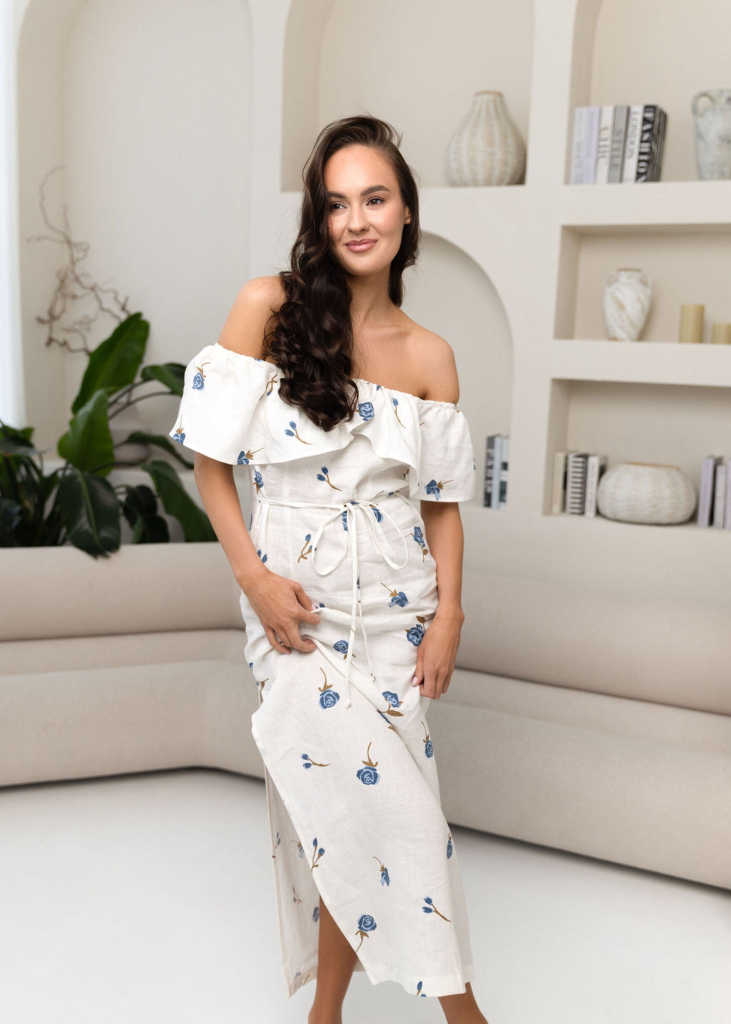 Woman wearing a white off-shoulder dress with floral patterns in a modern living room.