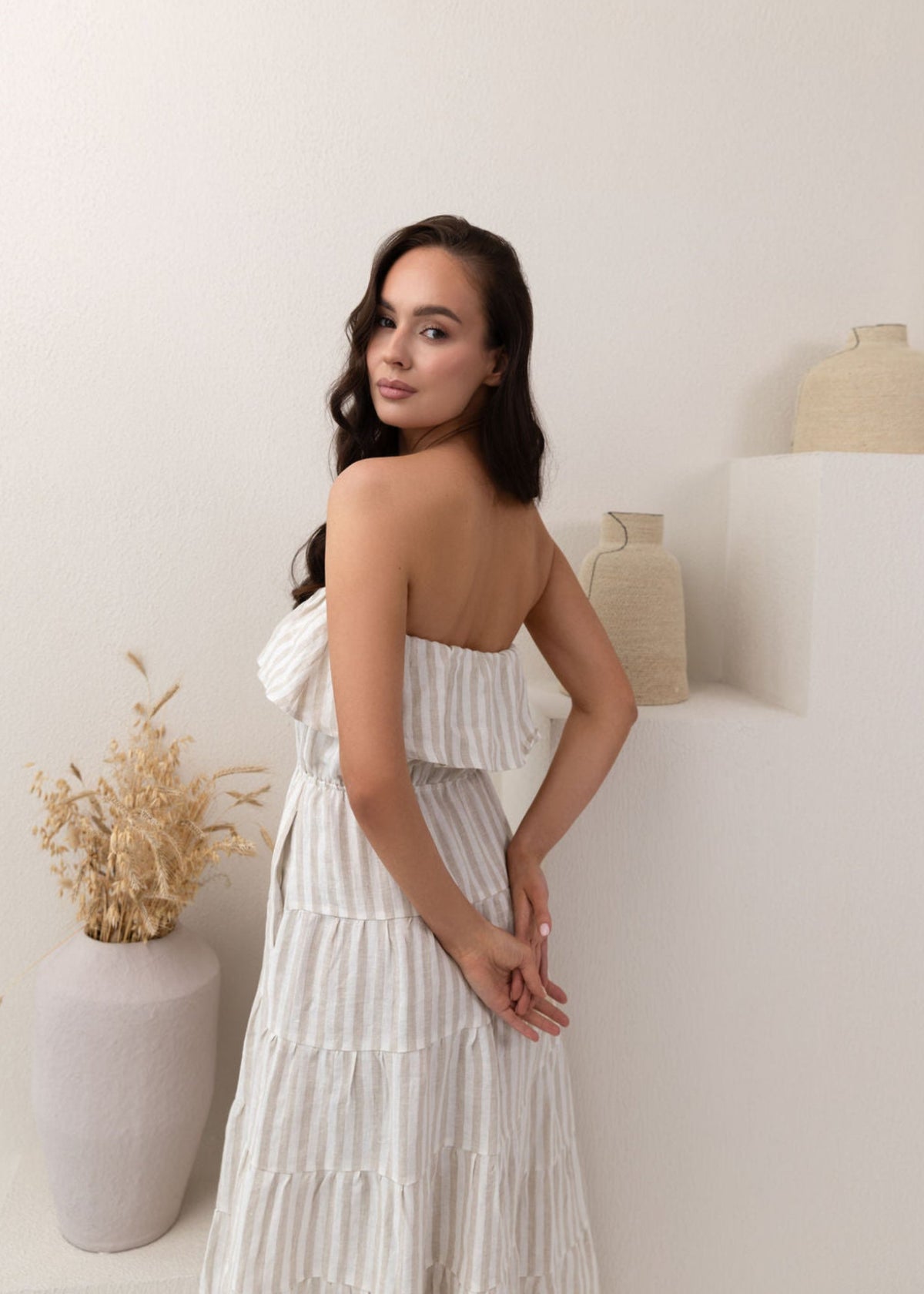 Woman in a white linen dress standing in a minimalistic room with a vase and plant.