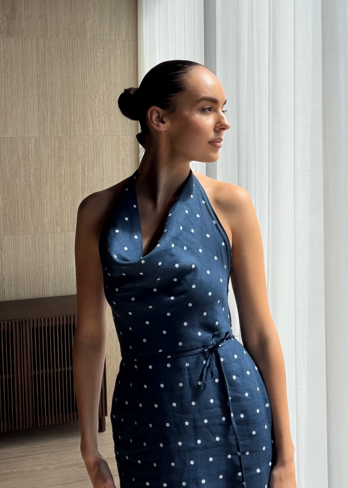 A woman wearing a navy blue halter-neck dress with a white polka dot pattern, standing in a modern interior setting with a textured beige wall and white curtains.
