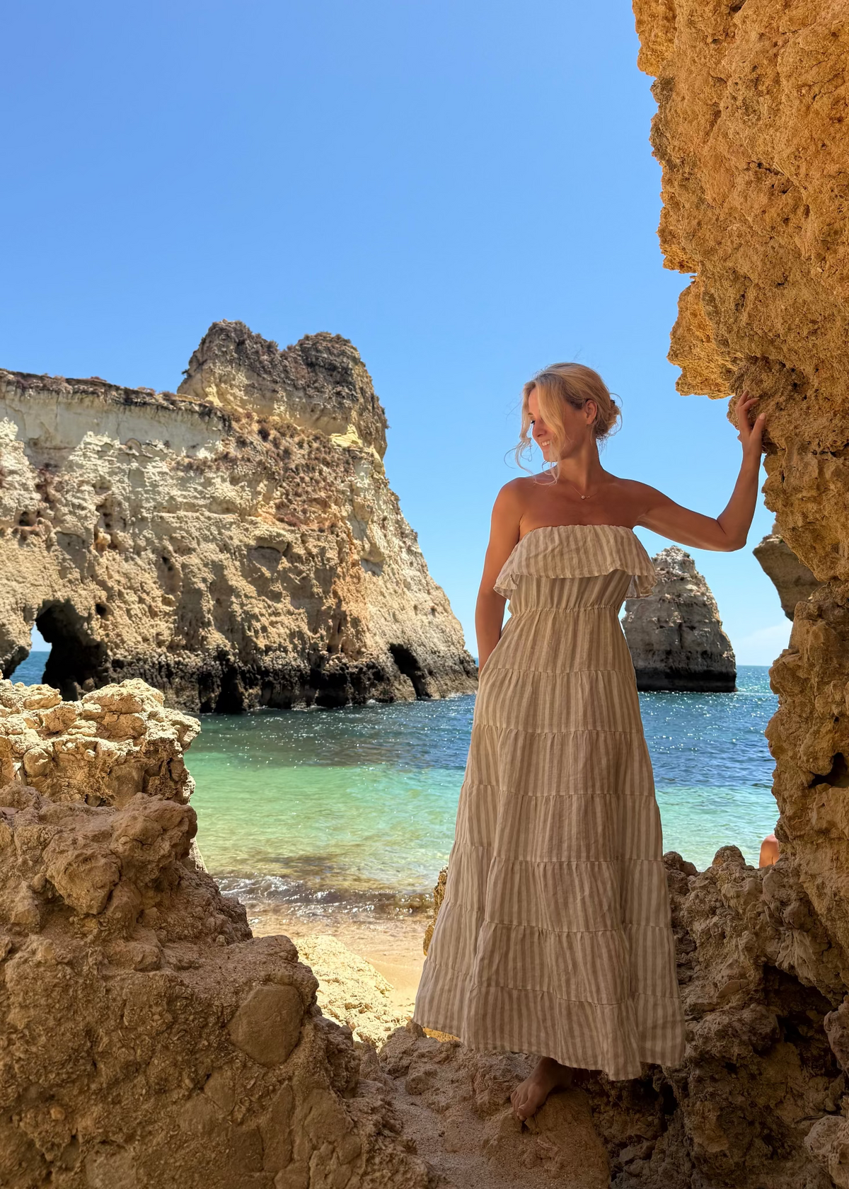 Woman in a Bohomey linen maxi dress standing between two rocky formations with a scenic ocean view.