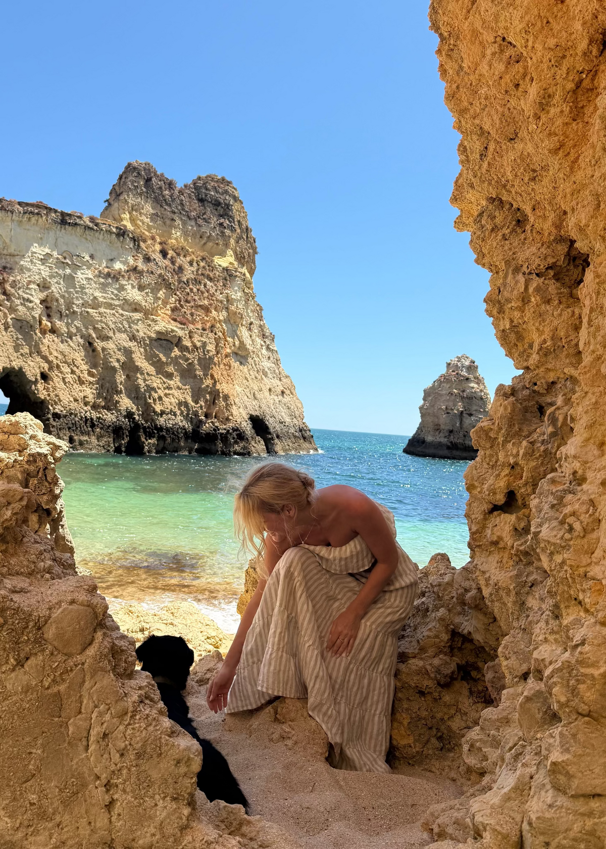 Woman in a Bohomey striped linen maxi dress sitting on a sandy beach with rocky cliffs and clear blue water.
