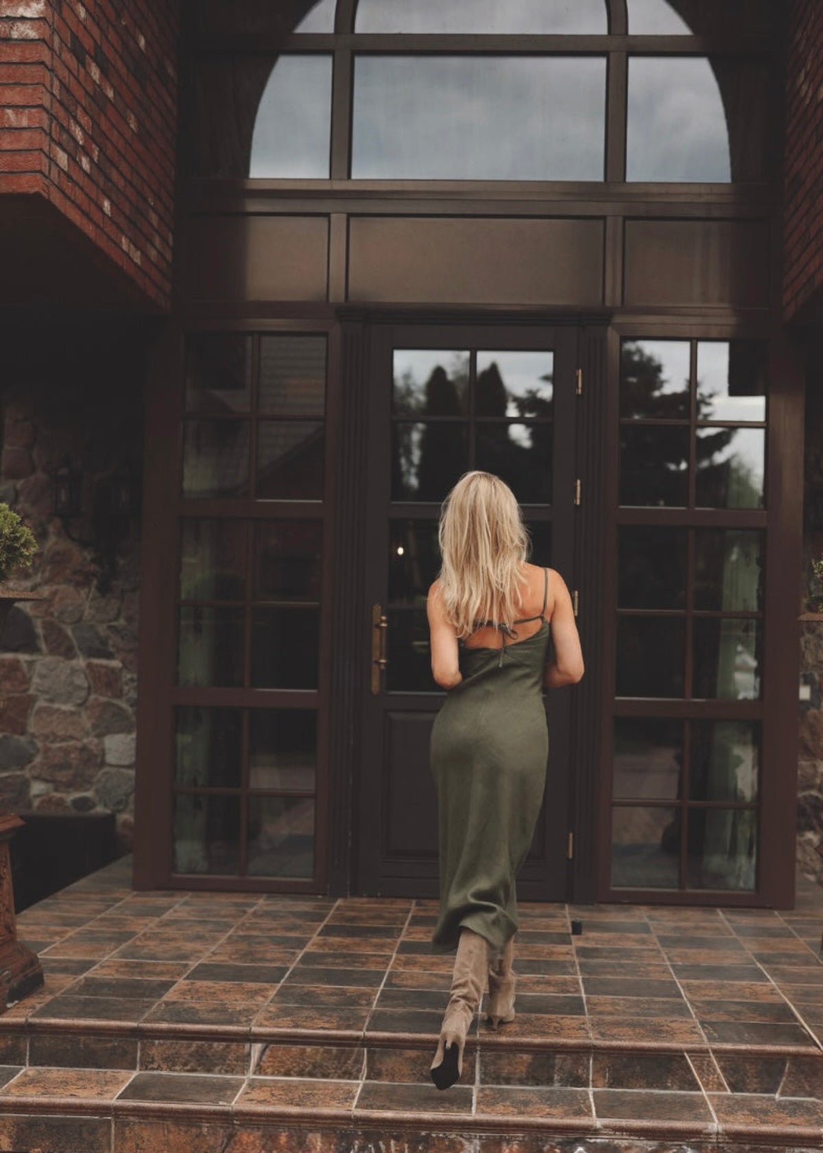 Woman in a Bohomey moss green linen slip dress walking towards a large glass door of a stone building.