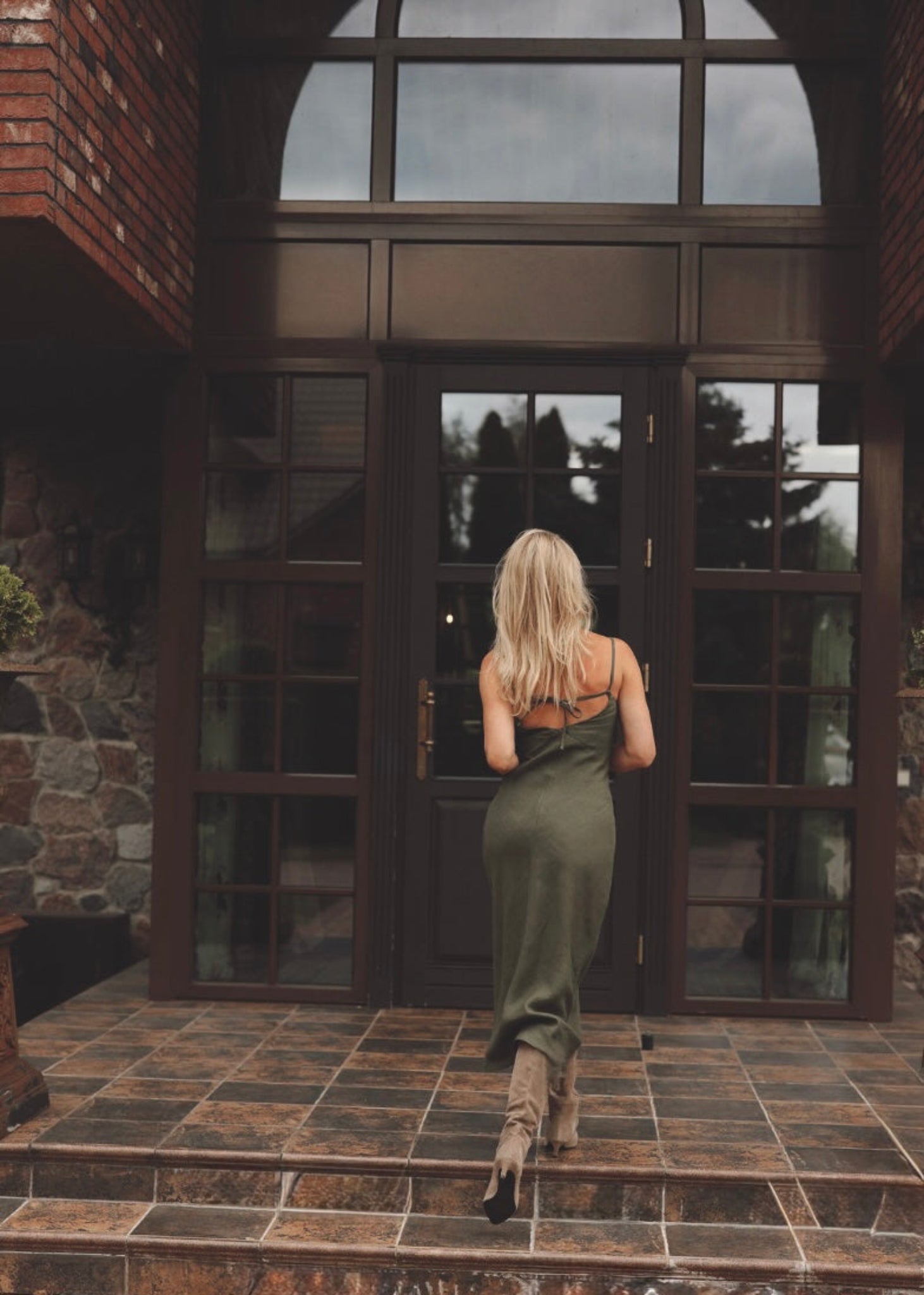 Woman in a Bohomey moss green linen slip dress standing in front of a large glass door with brick walls.