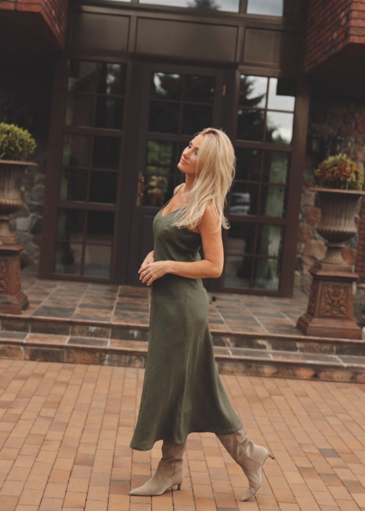 Woman in a Bohomey moss green linen slip dress standing in front of a brick building with large windows.
