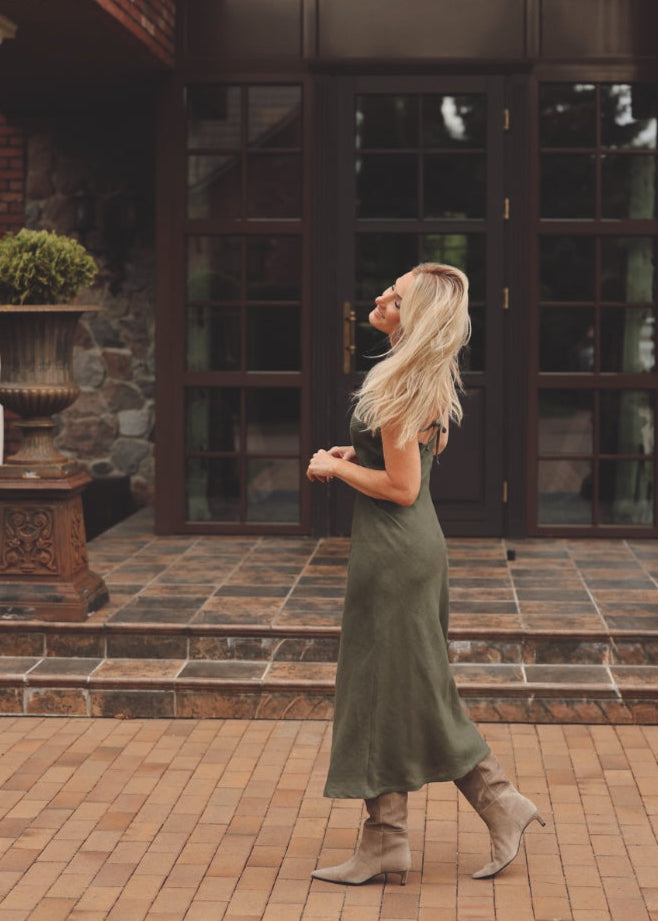 Woman in a Bohomey moss green linen slip dress standing in front of a large glass door with brick walls.