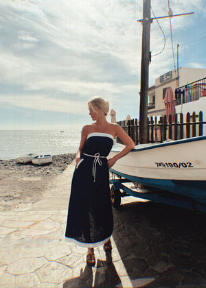 Woman in a strapless linen dress standing by a boat on a beach with a cloudy sky.