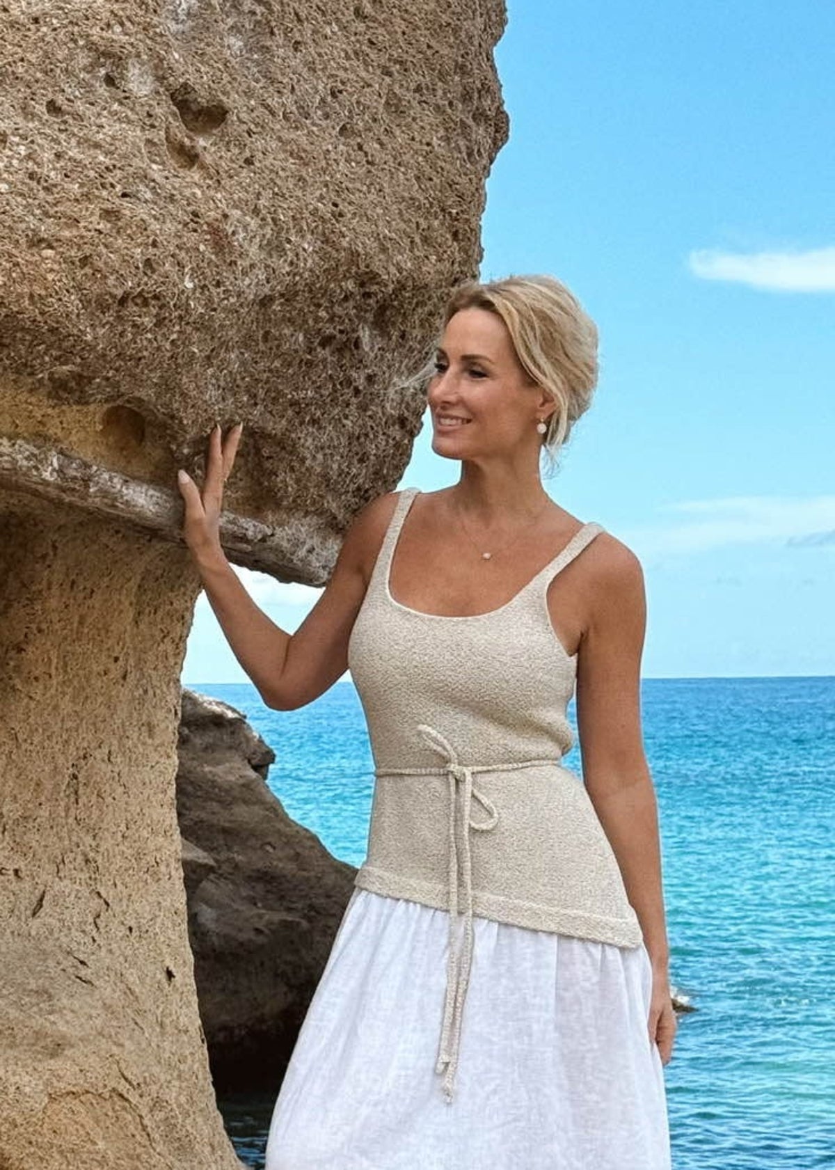 Woman in a Bohomey beige and white linen dress standing by a rocky coastline with blue water and sky.