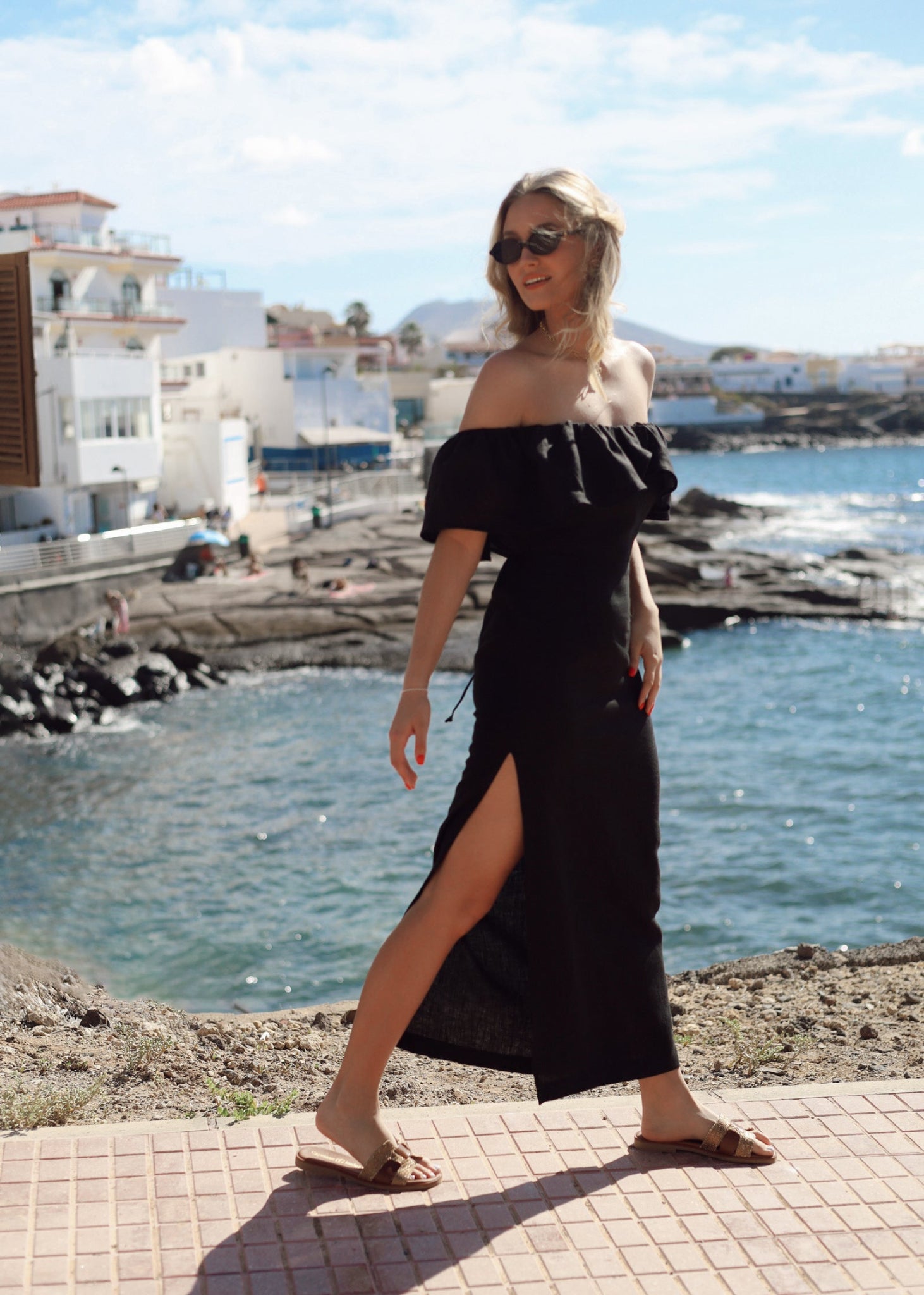 Woman in a black off-shoulder linen dress standing by a waterfront with buildings and water in the background.