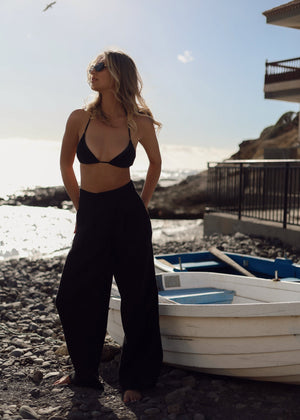 Woman in a black bikini and Bohomey linen pants standing on a rocky beach with boats and a clear sky.