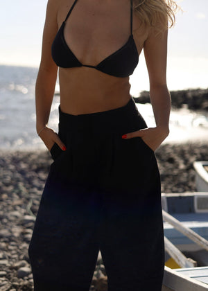 Woman wearing a black bikini top and Bohomey black linen pants on a beach.