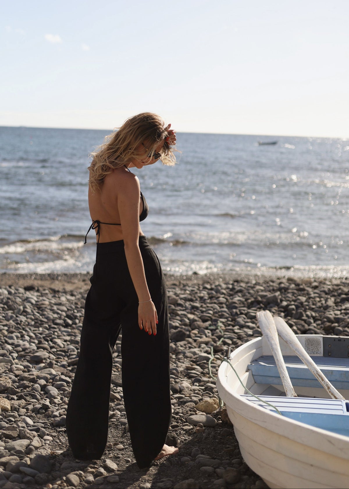 Woman wearing Bohomey black linen pants standing on a beach near a white boat with ocean in the background