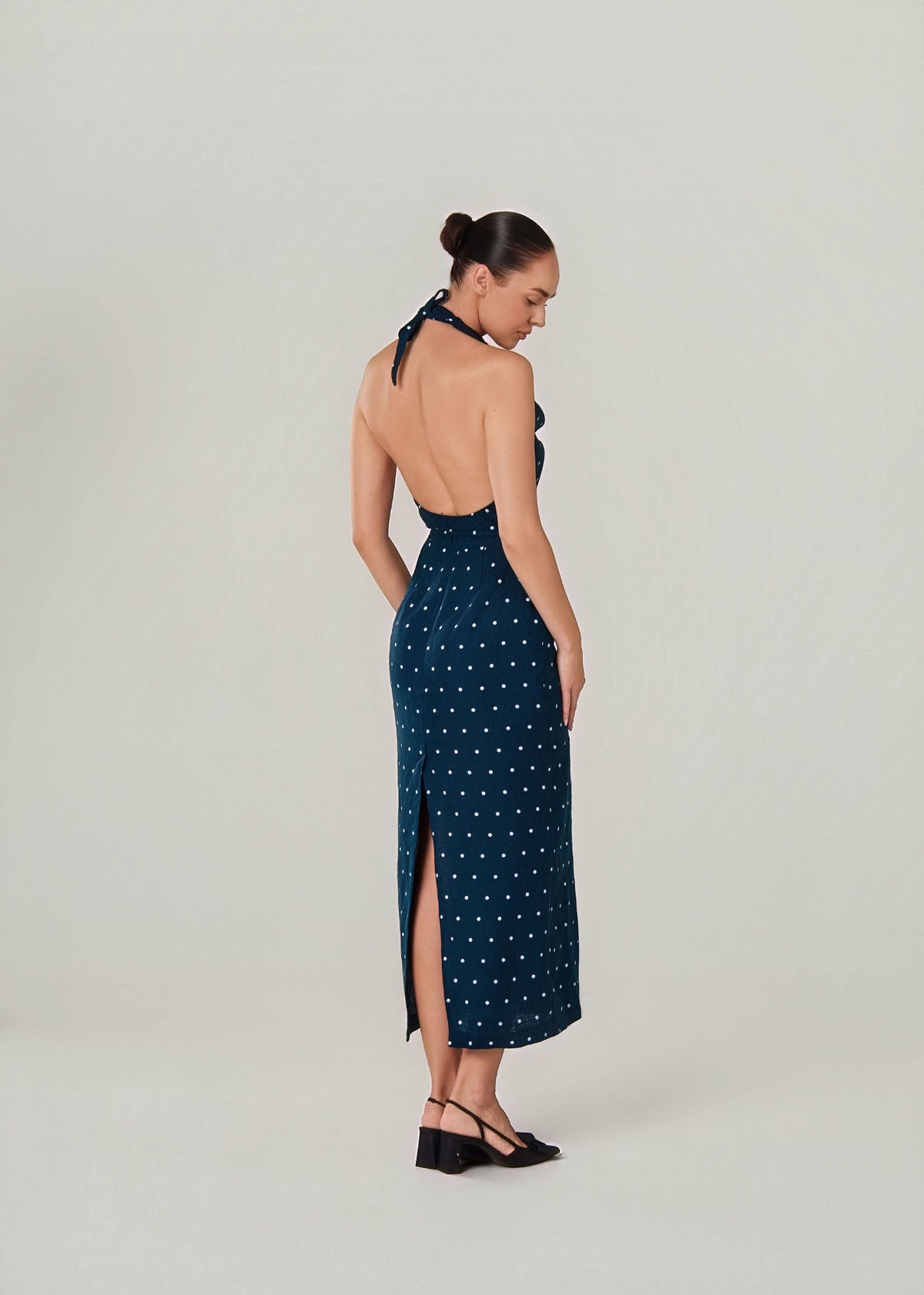 A woman modeling a dark teal halter-neck maxi dress with a white polka dot linen pattern, shown from the back against a light gray studio background.