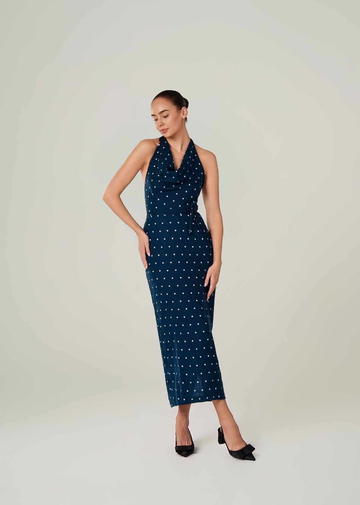 A woman modeling a sleeveless navy blue halter-neck midi dress with a white polka dot linen pattern, paired with black pointed-toe heels, against a light gray studio background.