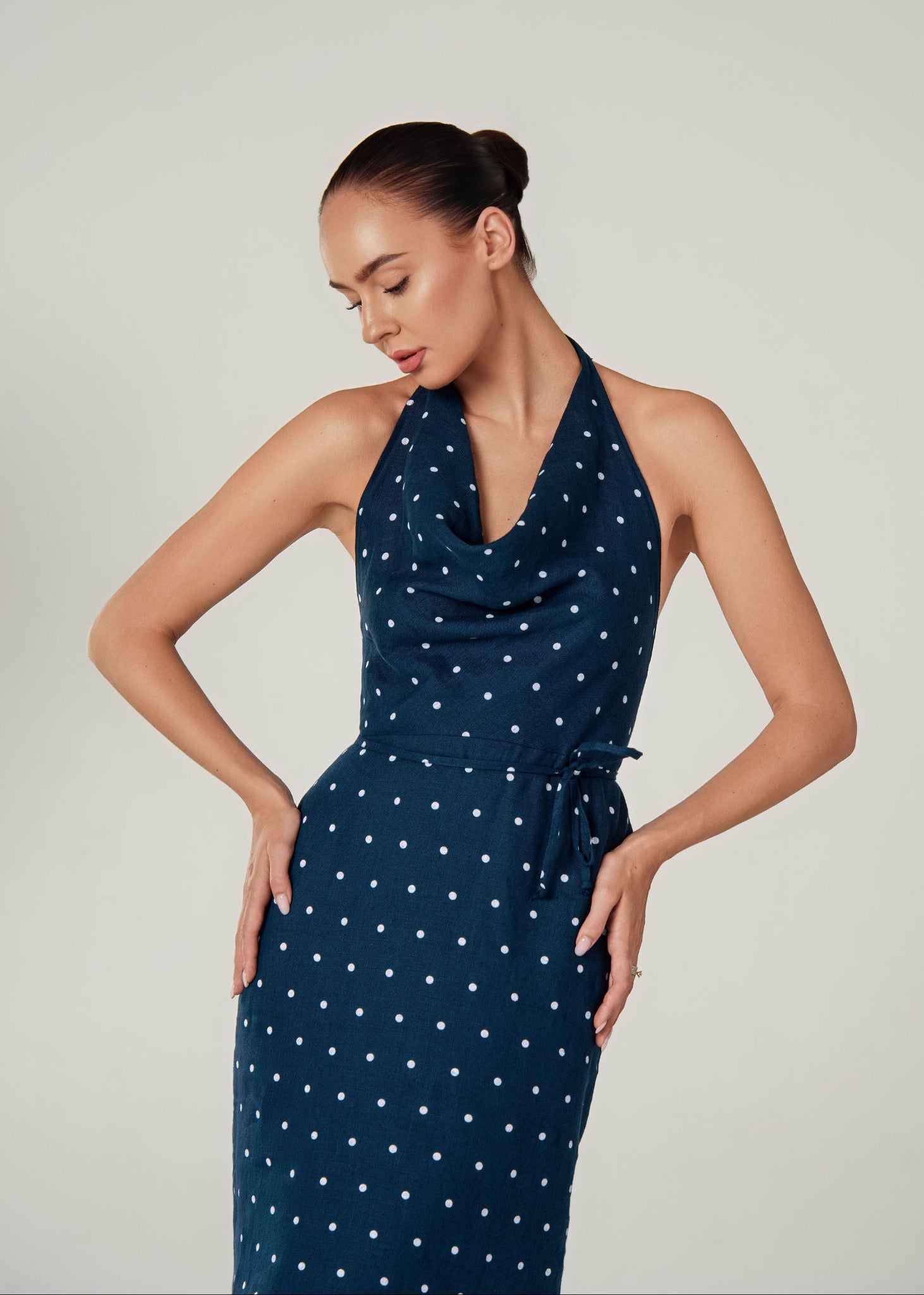 A woman modeling a navy blue halter-neck dress with a white polka dot linen pattern against a plain light gray studio background.