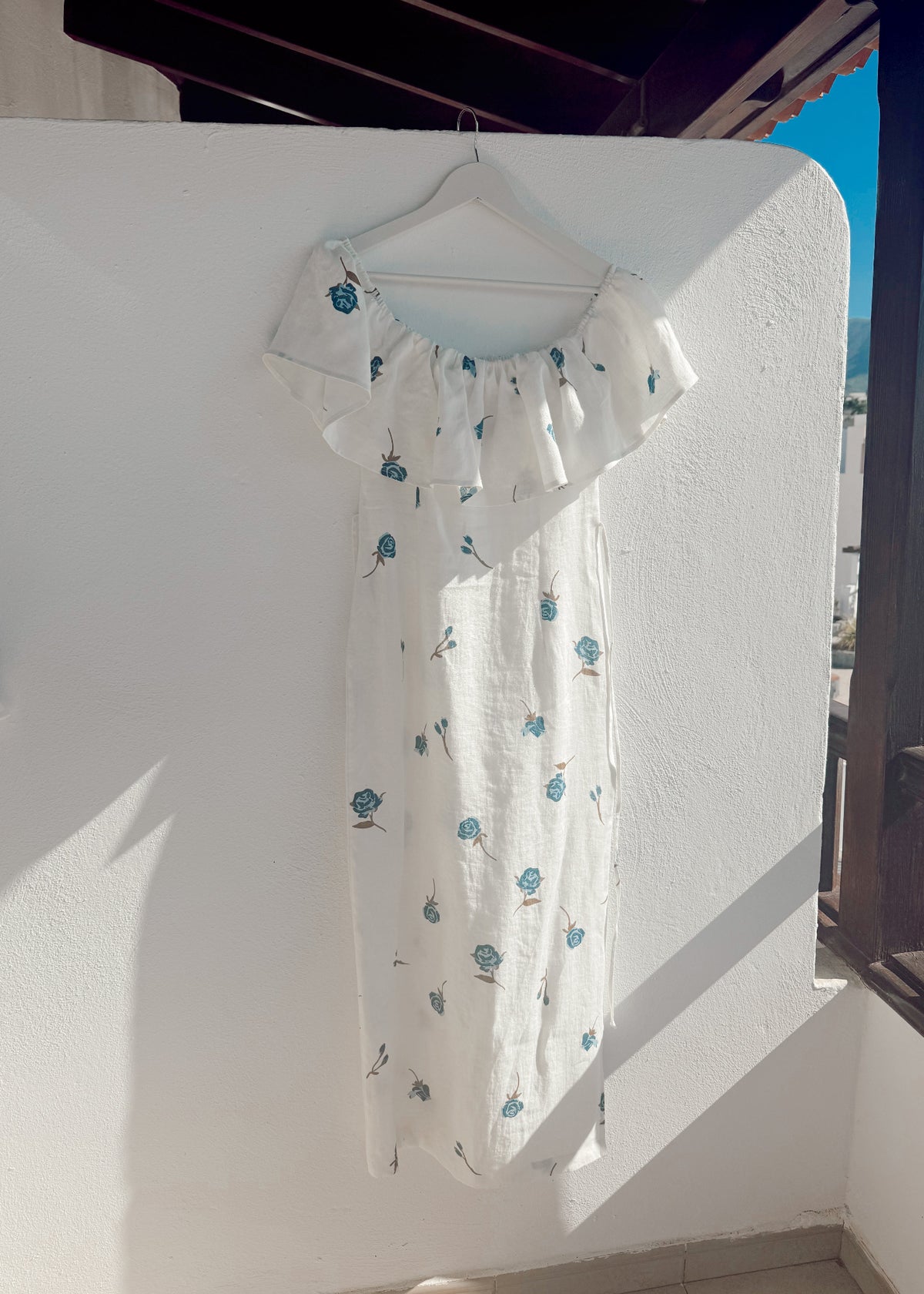 White linen dress with blue floral patterns on a hanger against a white wall.