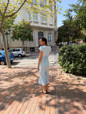 A woman wearing a light blue midi dress stands on a brick sidewalk in front of a building with trees and parked cars.