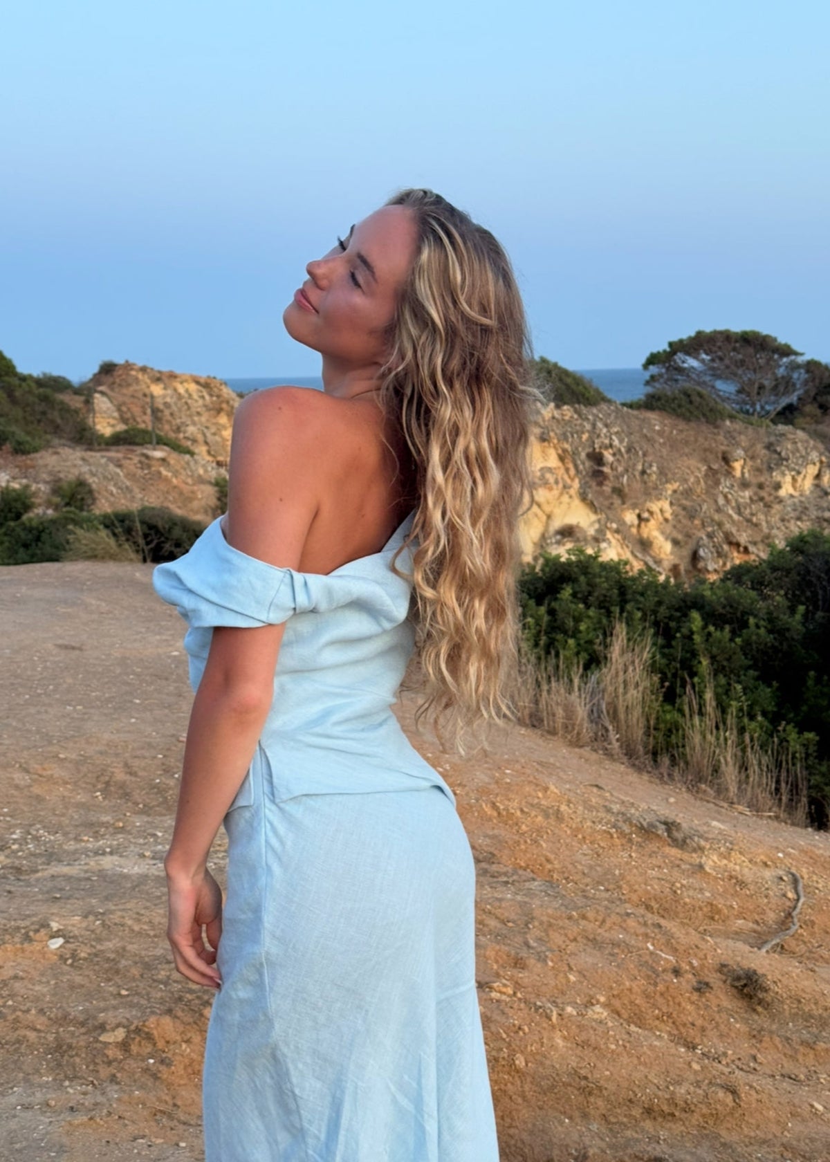 Woman in a BOHOMEY light blue linen dress standing on a scenic hillside with mountains and sky in the background.