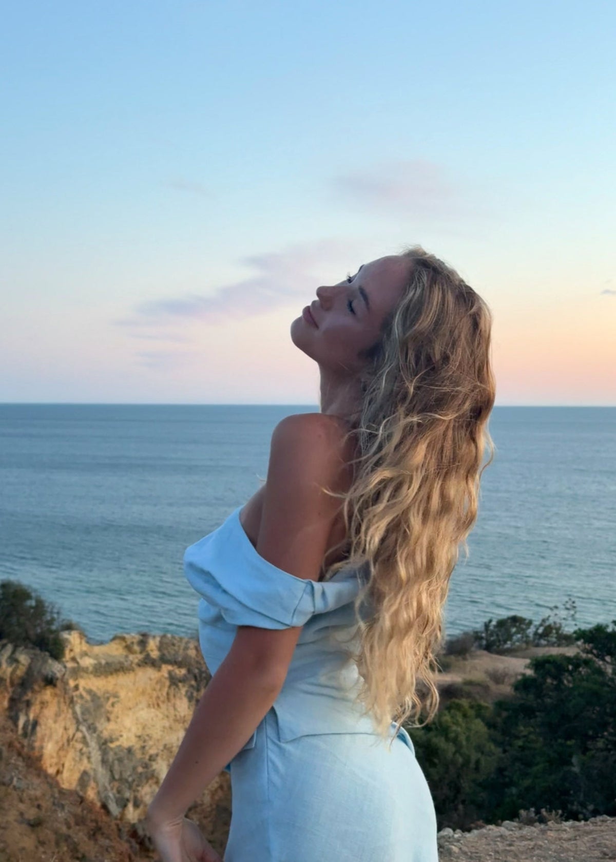 Woman in a light blue dress standing on a cliff overlooking the ocean at sunset.