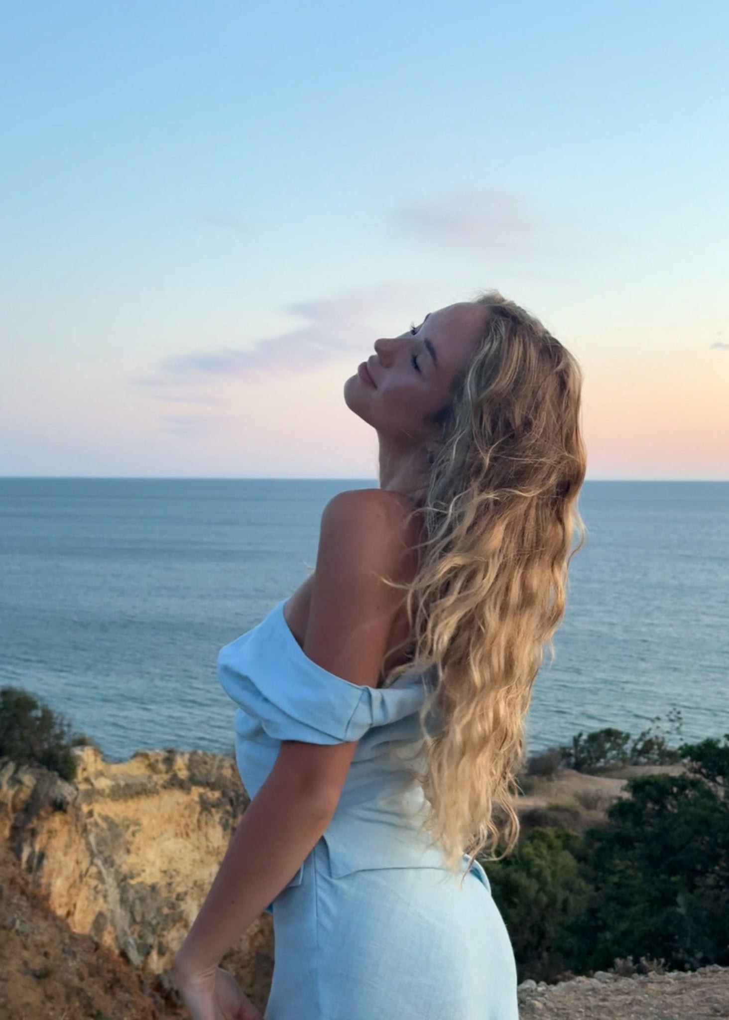 Woman in a light blue dress standing on a cliff overlooking the ocean at sunset.