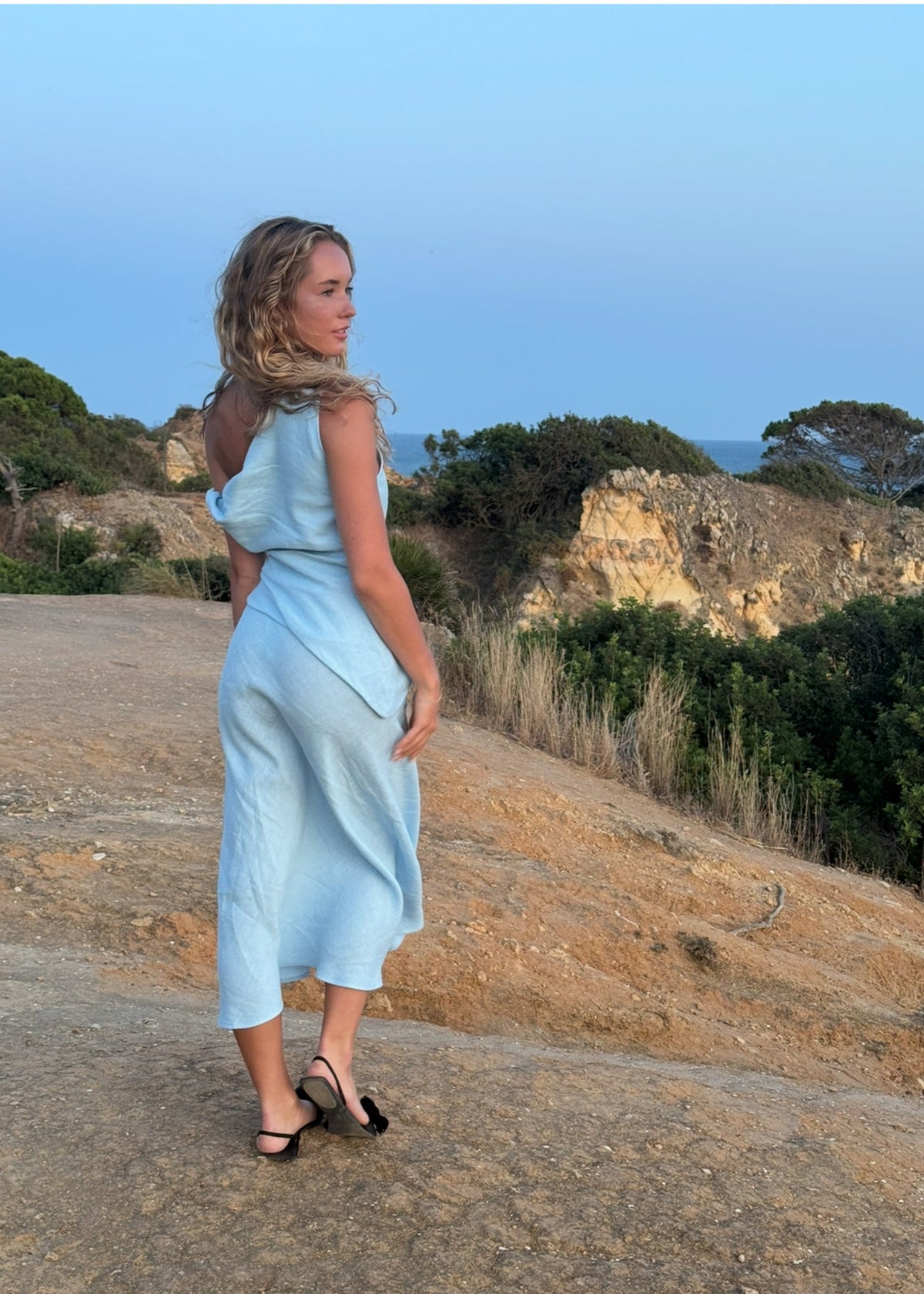 Woman in a BOHOMEY light blue linen dress standing on a scenic road with trees and sky in the background